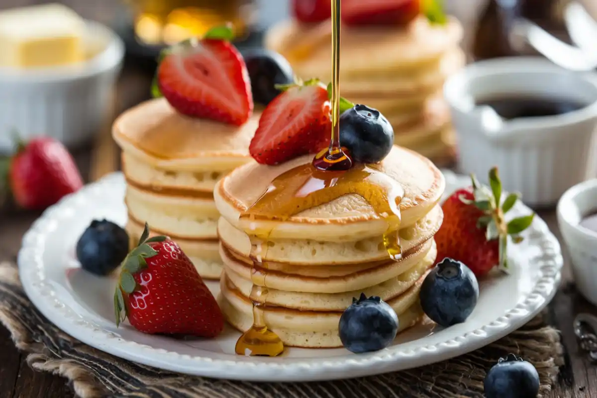 Homemade mini pancakes topped with fresh strawberries and blueberries, drizzled with syrup, on a breakfast table.