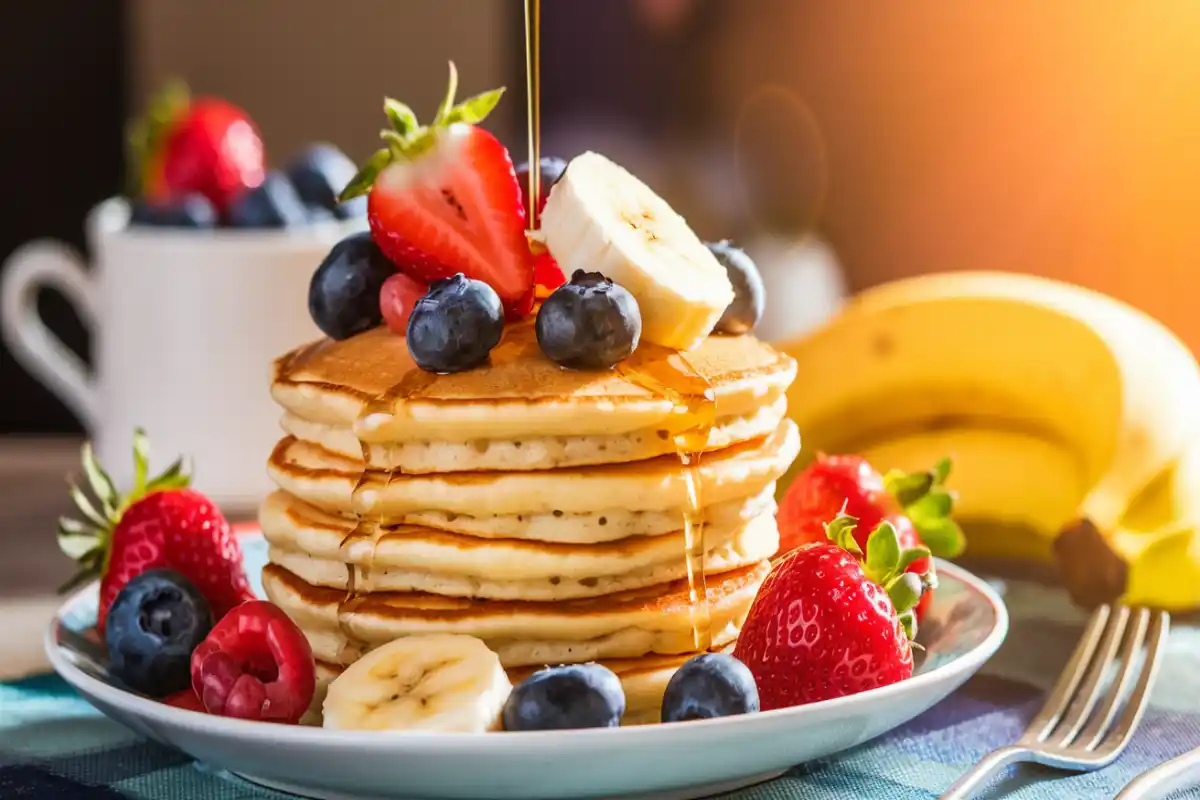 Stack of pancakes topped with fresh fruits like strawberries, blueberries, and bananas with honey drizzle.