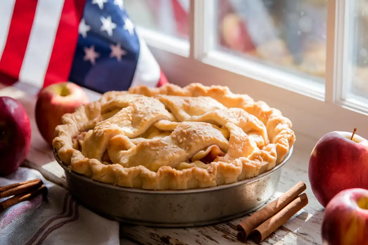 Golden apple pie on a wooden table with an American flag in the background, representing American culture and tradition.